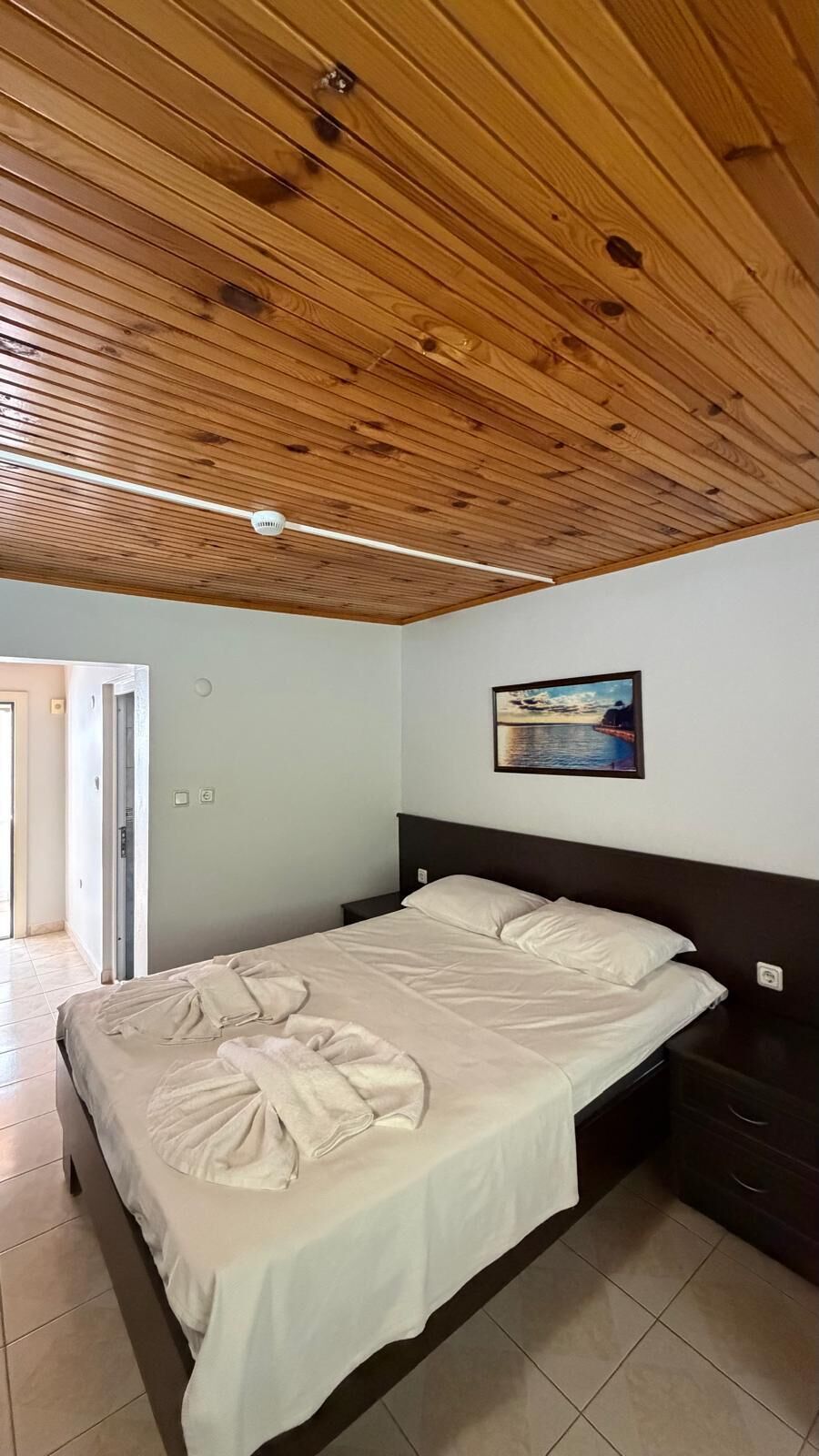 A bedroom with a wooden ceiling, a bed with a white bedspread, a nightstand, and a painting on the wall.