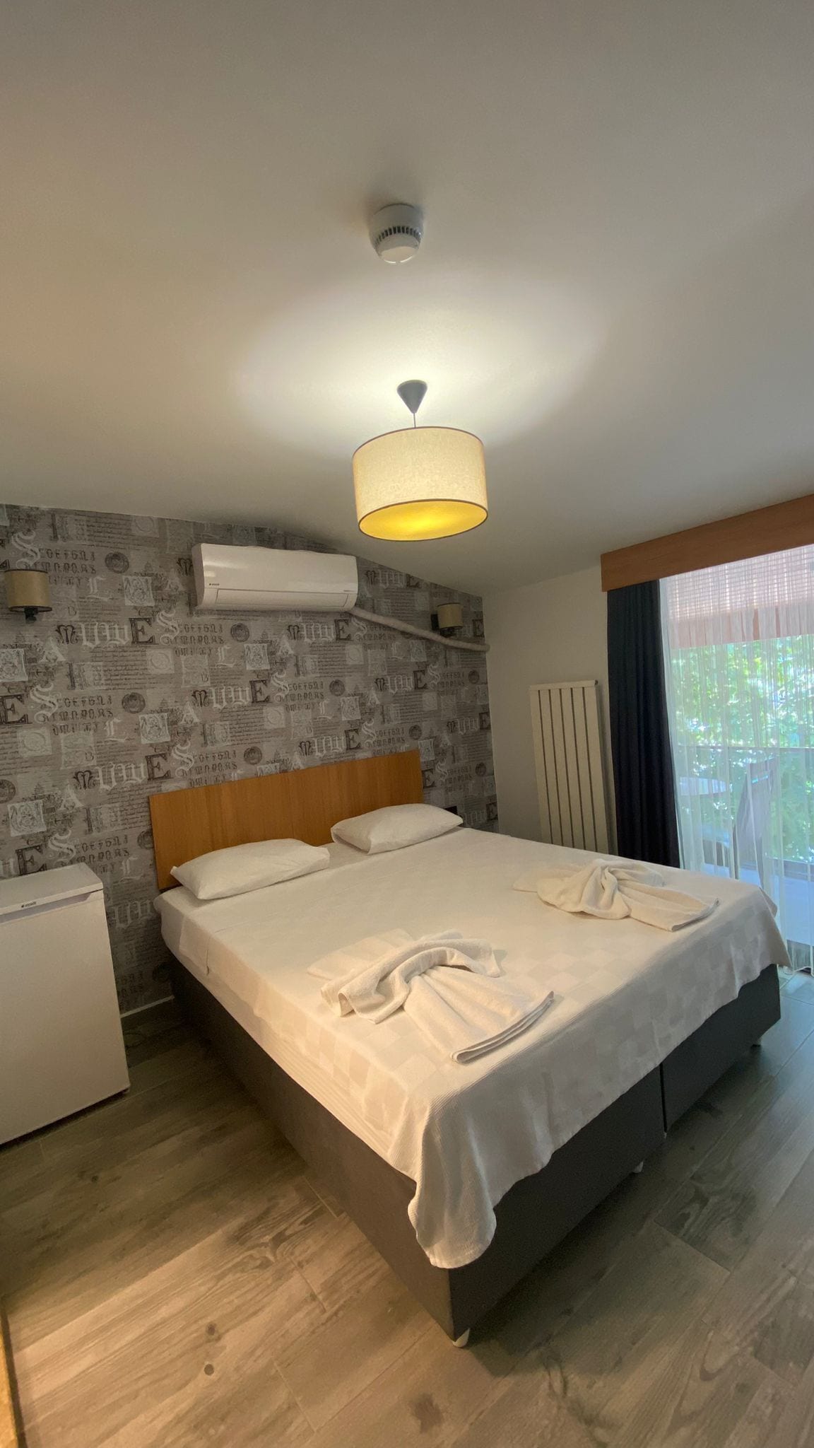 Een hotelkamer met een bed, nachtkastje, plafondlamp, radiator en een raam met gordijnen.