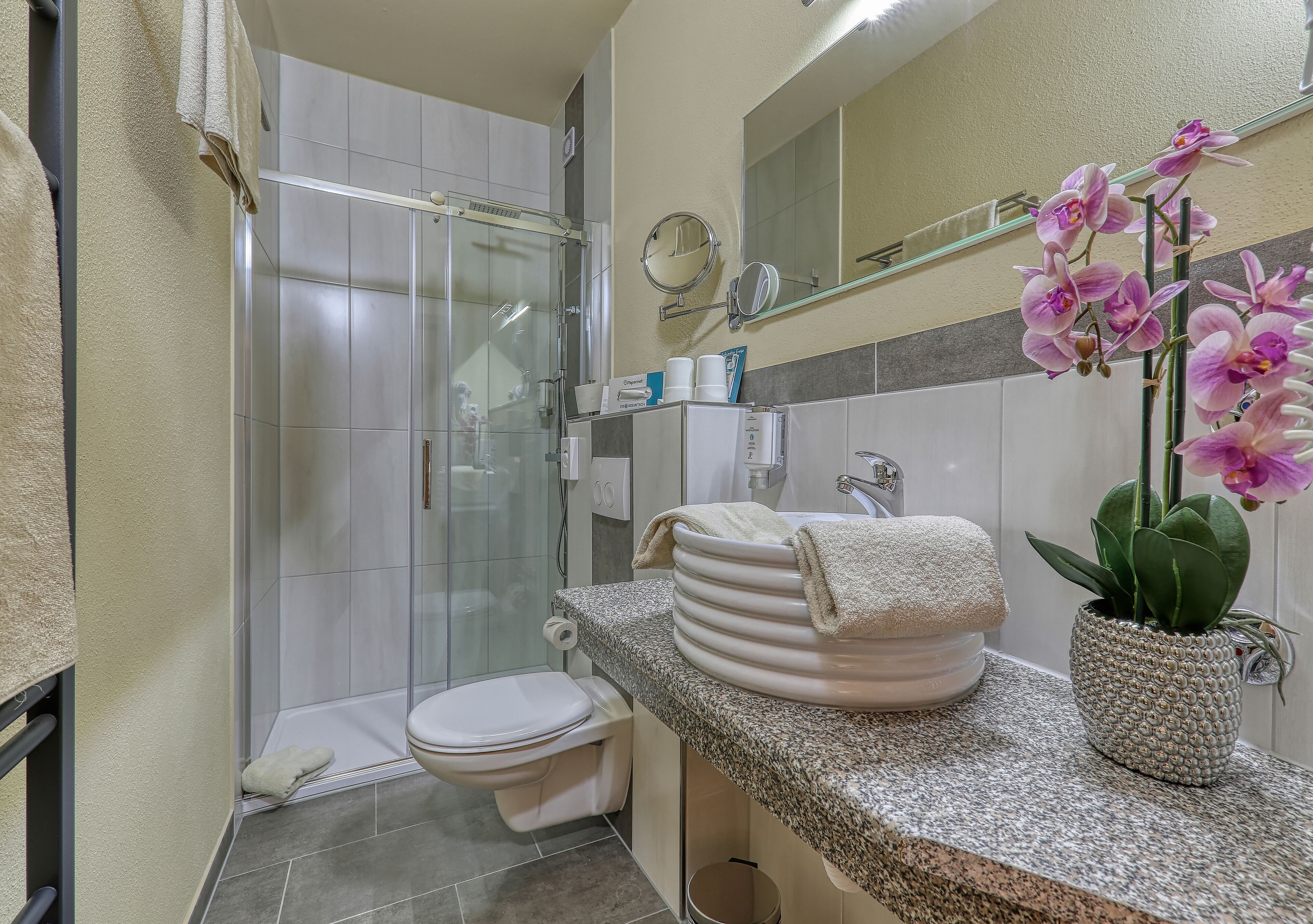 Een moderne badkamer met een douchecabine van glas, een toilet, een wastafel met een stap handdoeken en een potorkidee.