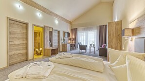 A hotel room with a large bed, a desk, a chair, and a small table. There is a door leading to a bathroom and a window with curtains. - Wellnesshotel Auszeit (Achslach)