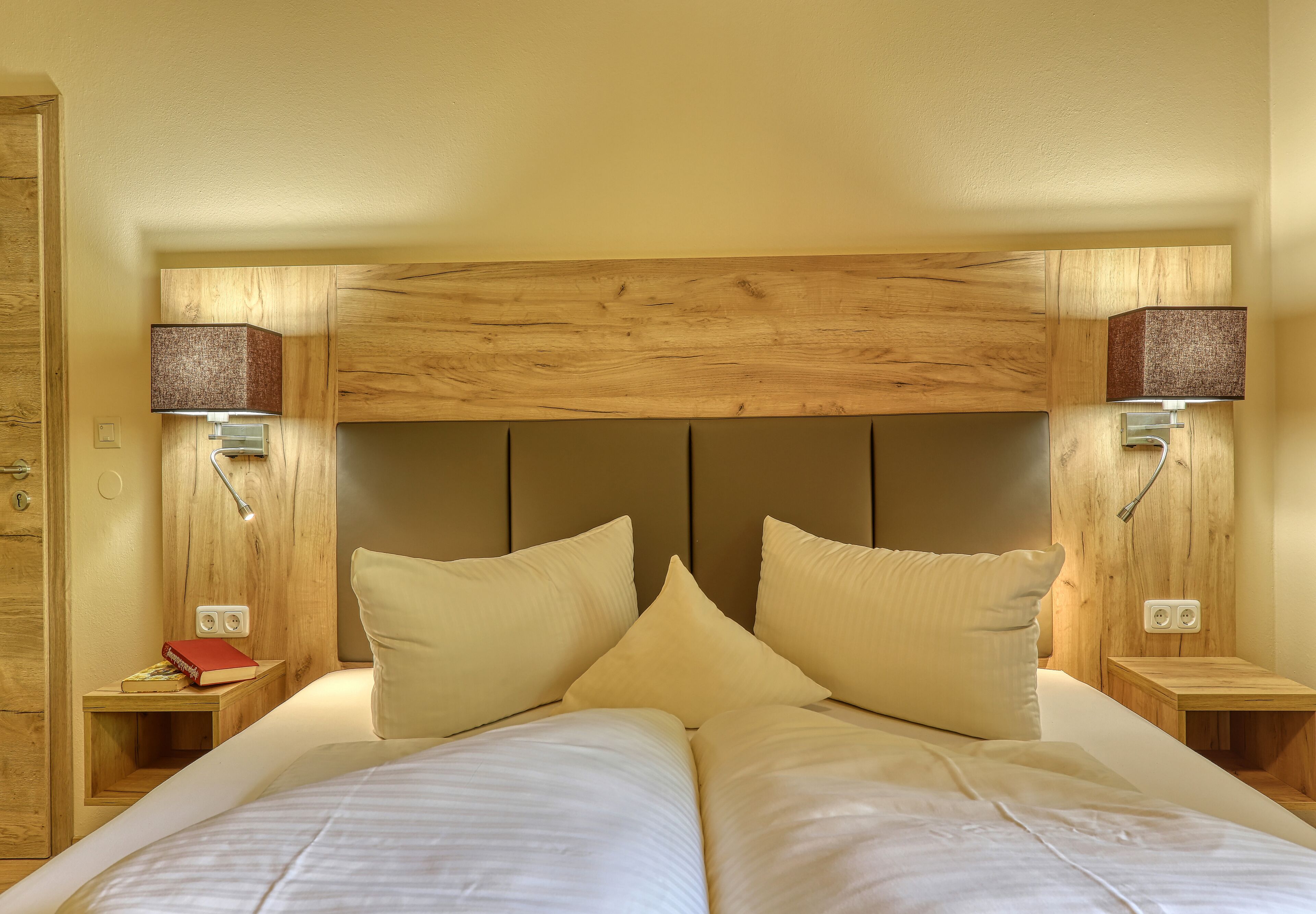 A double bed with two pillows, wooden headboard, bedside tables with lamps, and a small nightstand with a book.