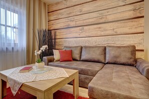A living room with a wooden wall, a brown sofa, a red cushion, a table with a book and a vase of flowers. - Wellnesshotel Auszeit (Achslach)
