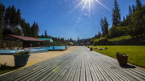 Eine Holzterrasse führt zu einem Schwimmbecken, im Hintergrund ein klarer, blauer Himmel und Bäume.