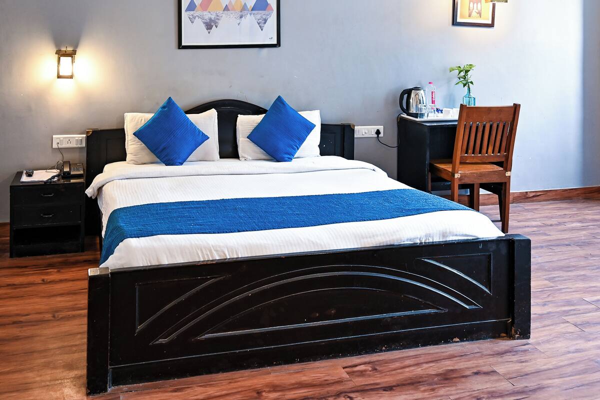 Een net opgemaakt bed met blauw-witte beddengoed, een zwart hoofdbord en een blauwe plaid. Rechts staat een bureau met een stoel, aan de muur hangt een ingelijst schilderij en een wandlamp.