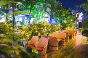 Un espacio al aire libre con sillas y mesas de mimbre, rodeado de plantas en macetas y palmeras, con un edificio al fondo.