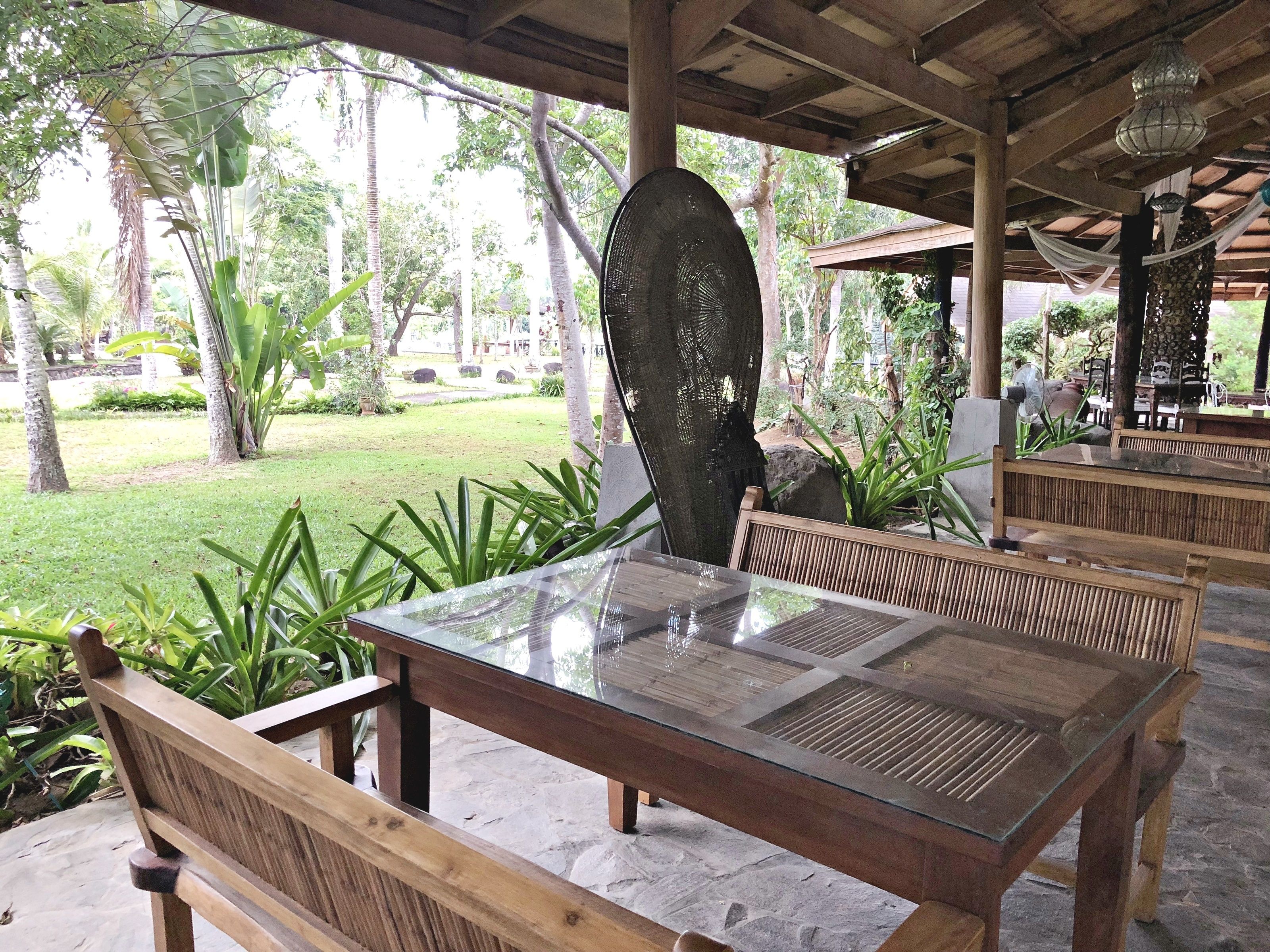 A covered outdoor seating area with wooden benches and a glass table, surrounded by lush greenery and a stone-paved floor.