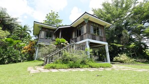 A two-story wooden house with a balcony, surrounded by lush greenery and a clear sky.