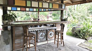 A rustic outdoor bar area with wooden chairs, a wooden counter, and a view of lush greenery.