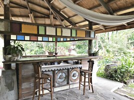 A rustic outdoor bar area with wooden chairs, a wooden counter, and a view of lush greenery.
