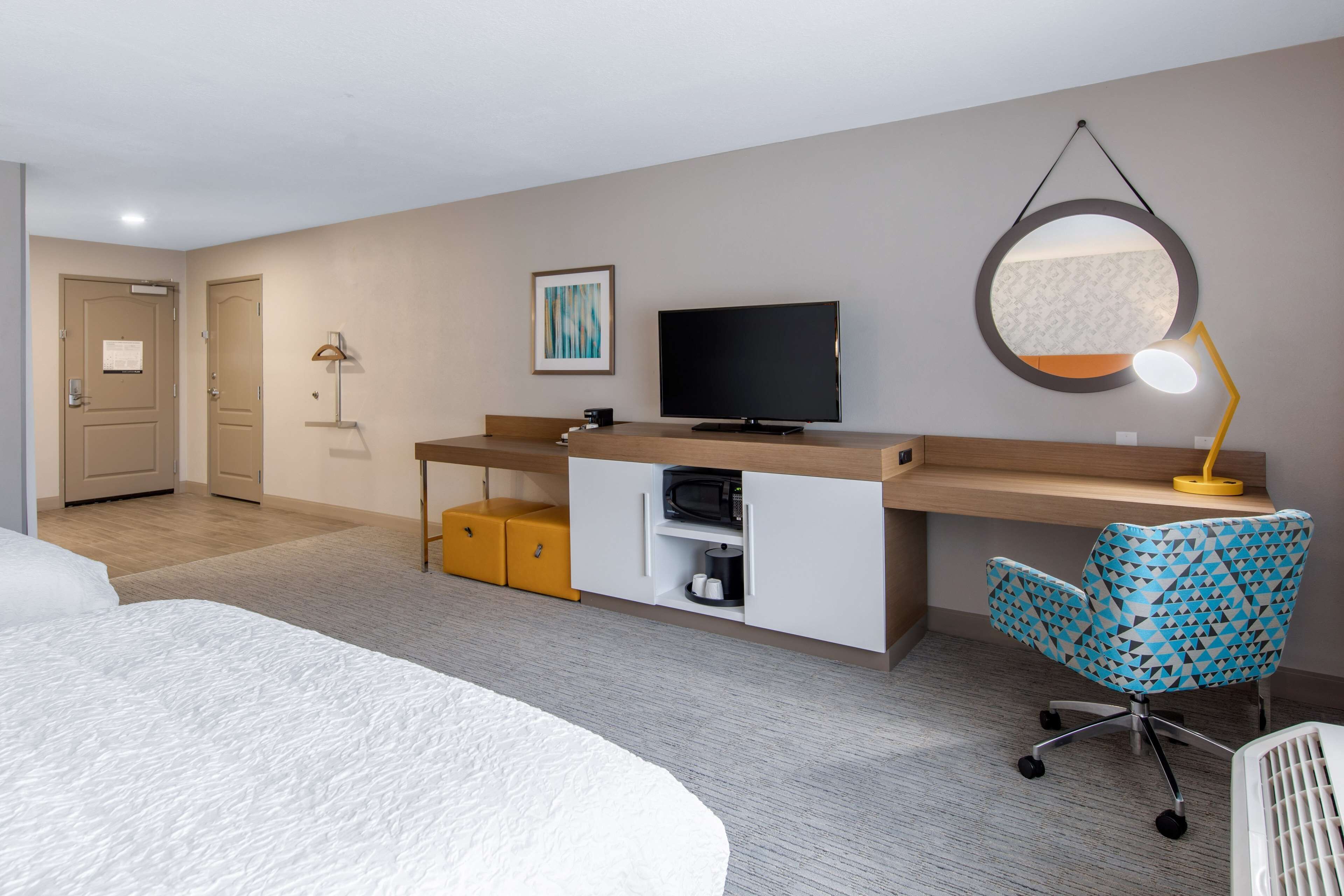 A hotel room with a bed, a desk with a TV, a desk lamp, and a chair.