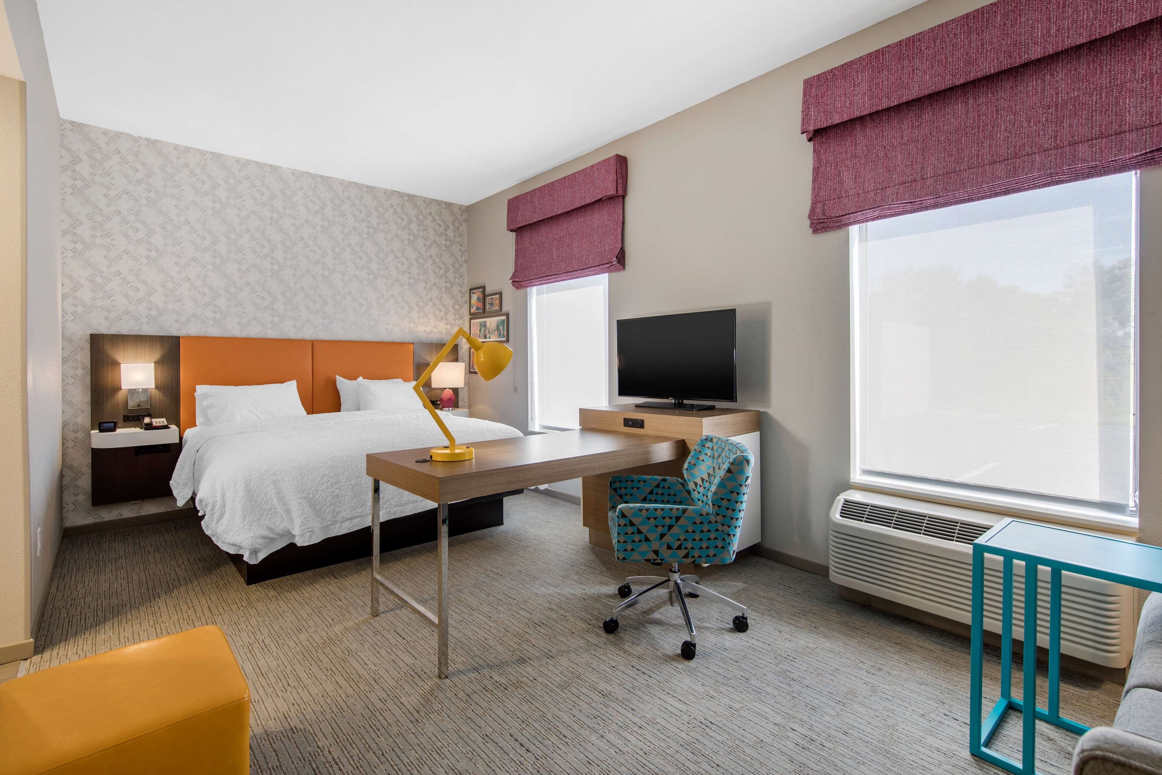 Habitación de hotel con cama, escritorio, silla, televisor y ventanas amplias.