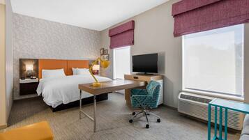 Habitación de hotel con cama, escritorio, silla, televisor y ventanas amplias.