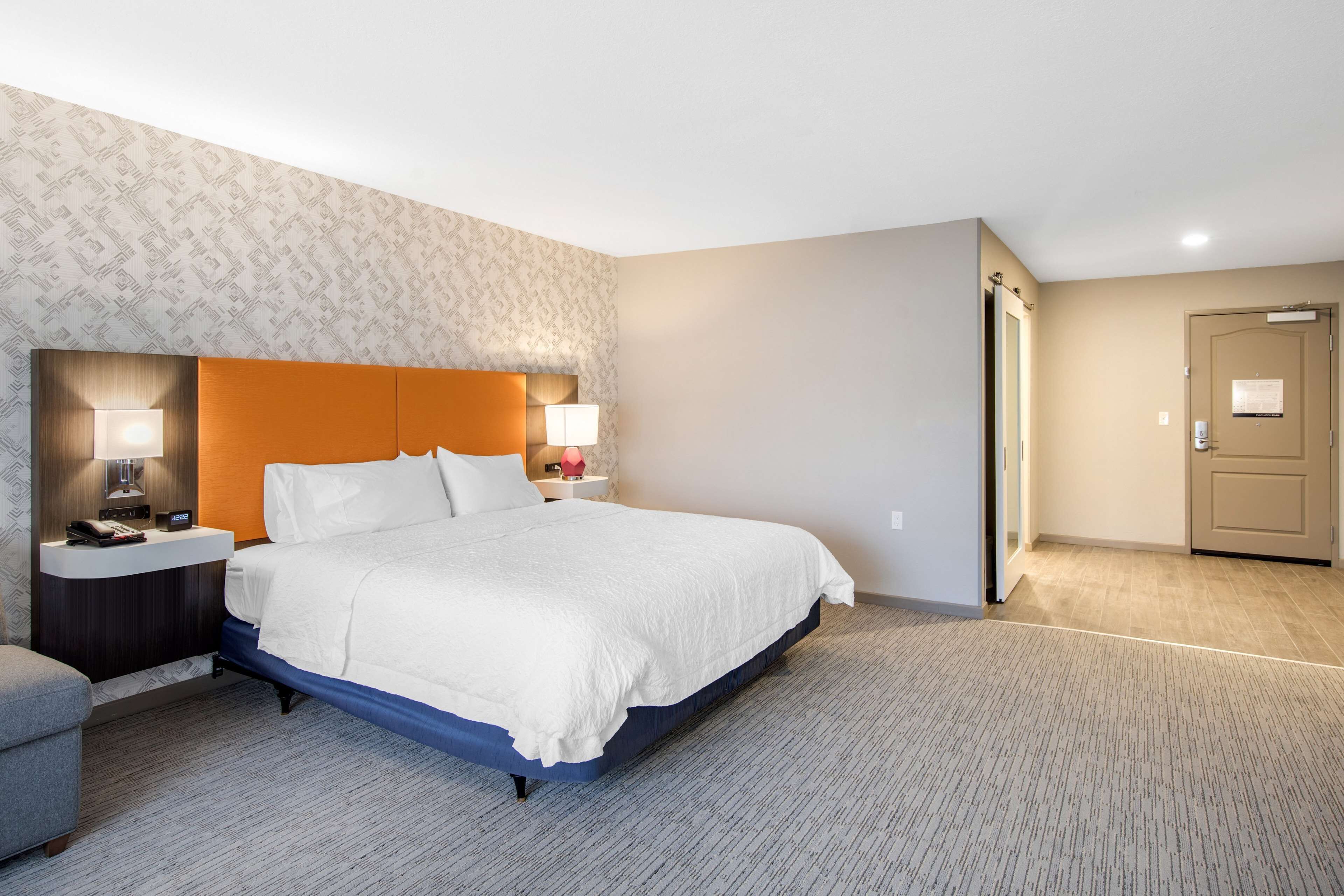 A hotel room with a large bed, two bedside tables with lamps, a small armchair, and a door.