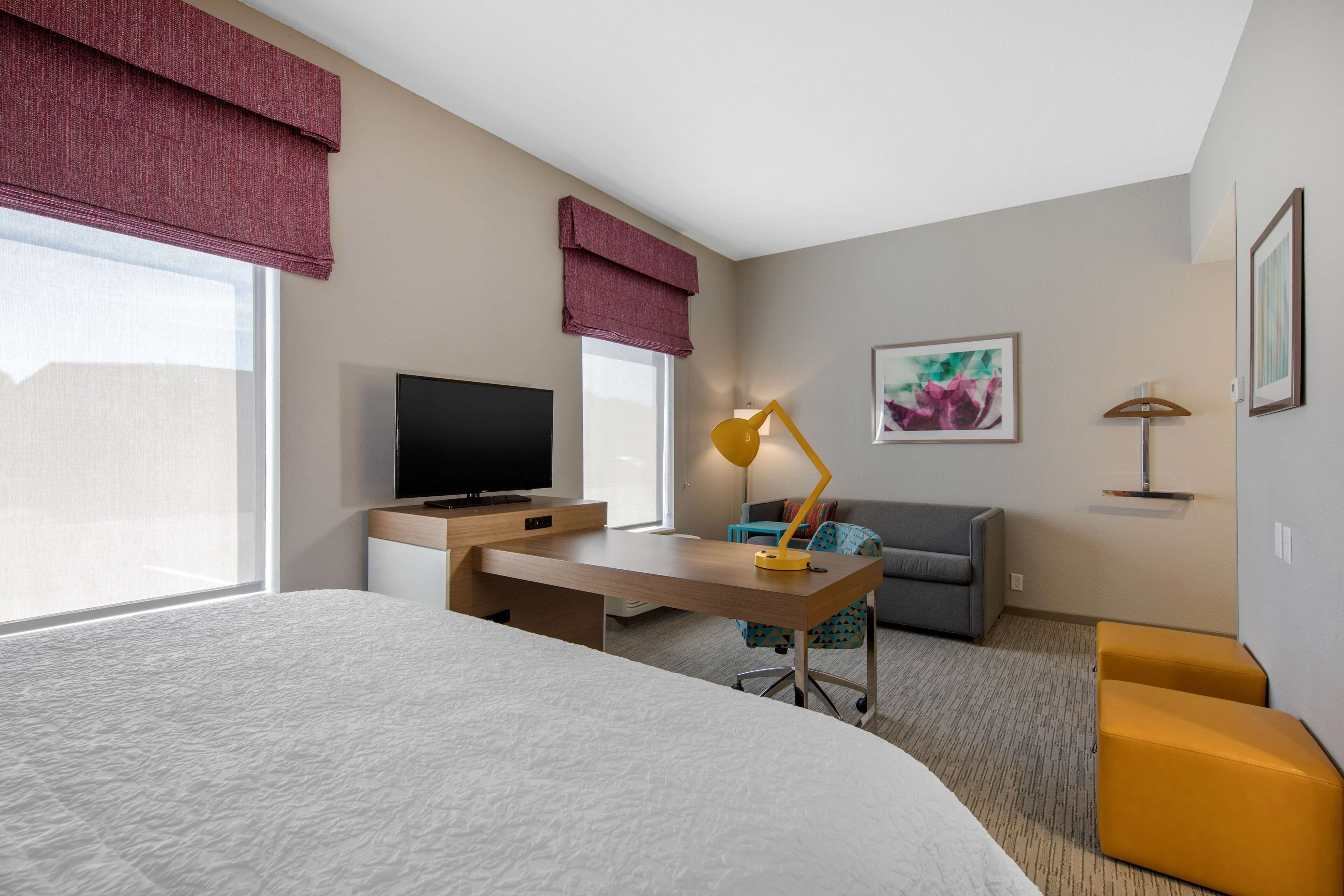 A hotel room with a bed, a desk with a lamp, a television, a sofa, and a window with curtains.