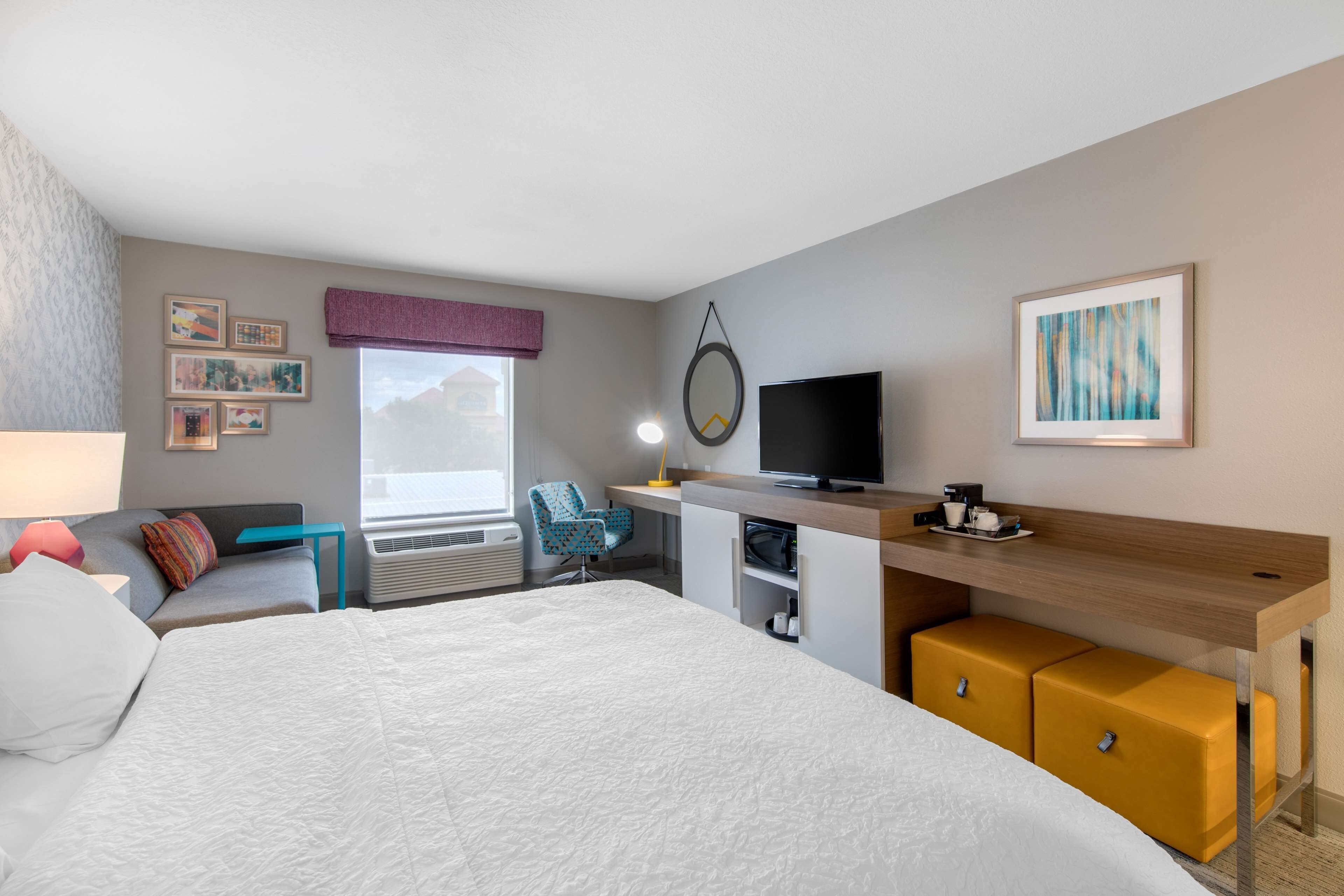 A modern hotel room with a large bed, a desk with a flat-screen TV, a chair, and a small seating area with a window view.