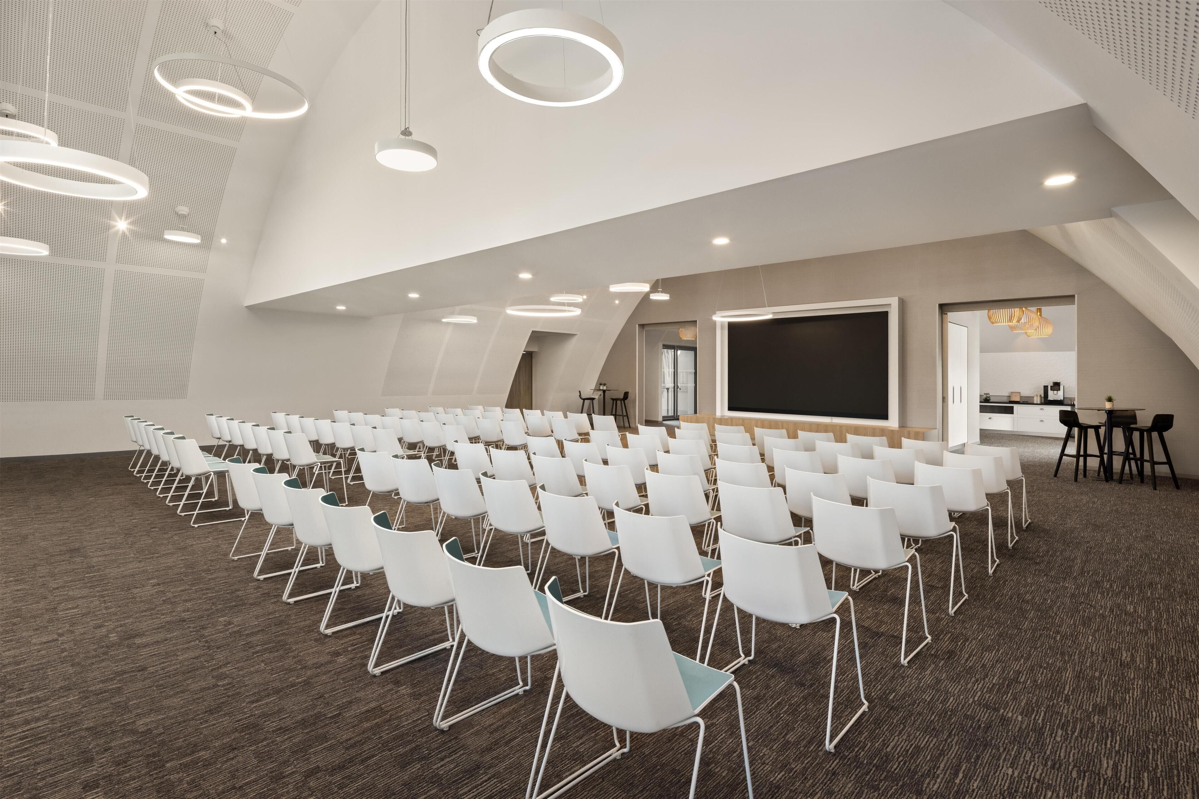 A conference room with white chairs arranged in rows, a large screen, and modern lighting fixtures.