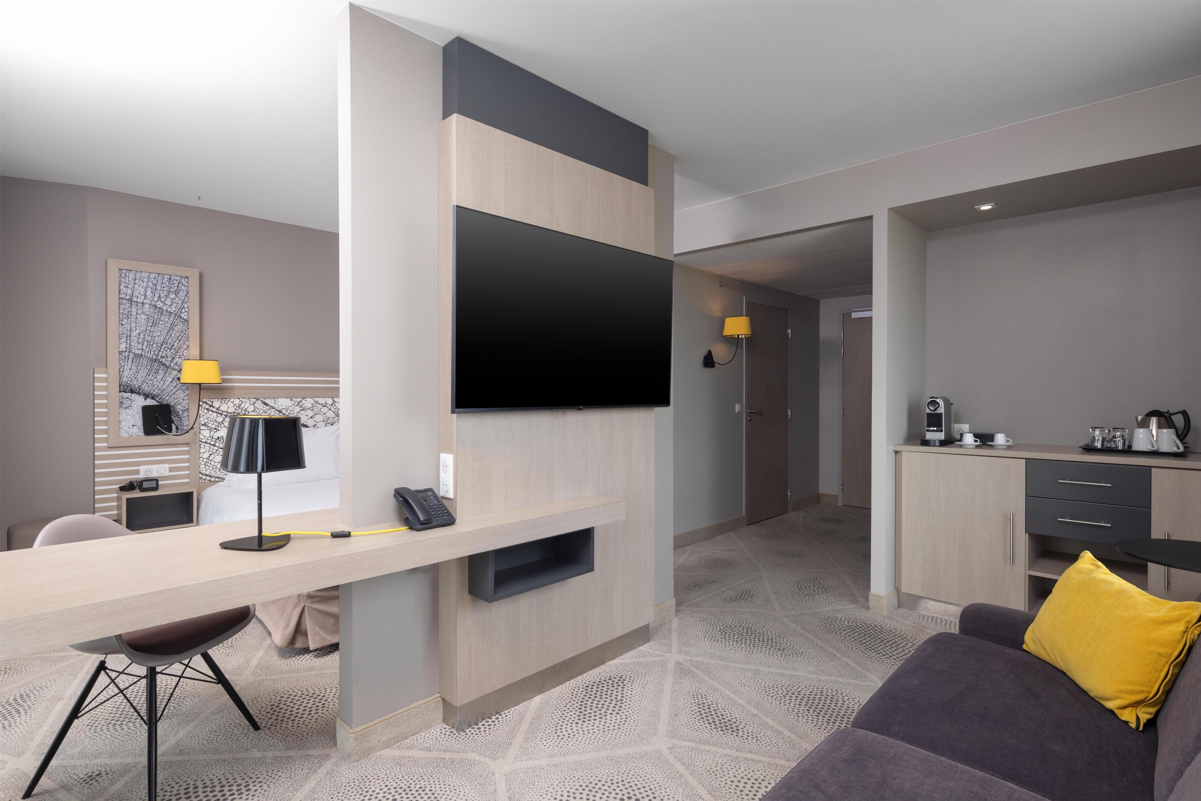 A modern hotel room with a flat-screen TV mounted on the wall, a desk with a lamp, a sofa with a yellow pillow, and a kitchenette area.
