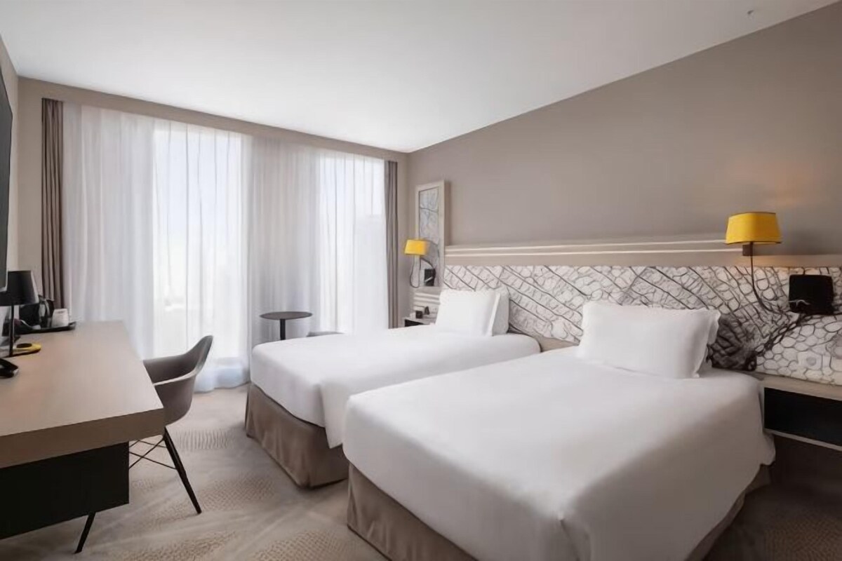 Une chambre d’hôtel avec deux lits, un bureau, une chaise, une lampe et une vue sur l’extérieur.
