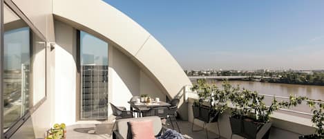 A modern balcony with a curved white wall, a dining area with chairs and a table, and potted plants.