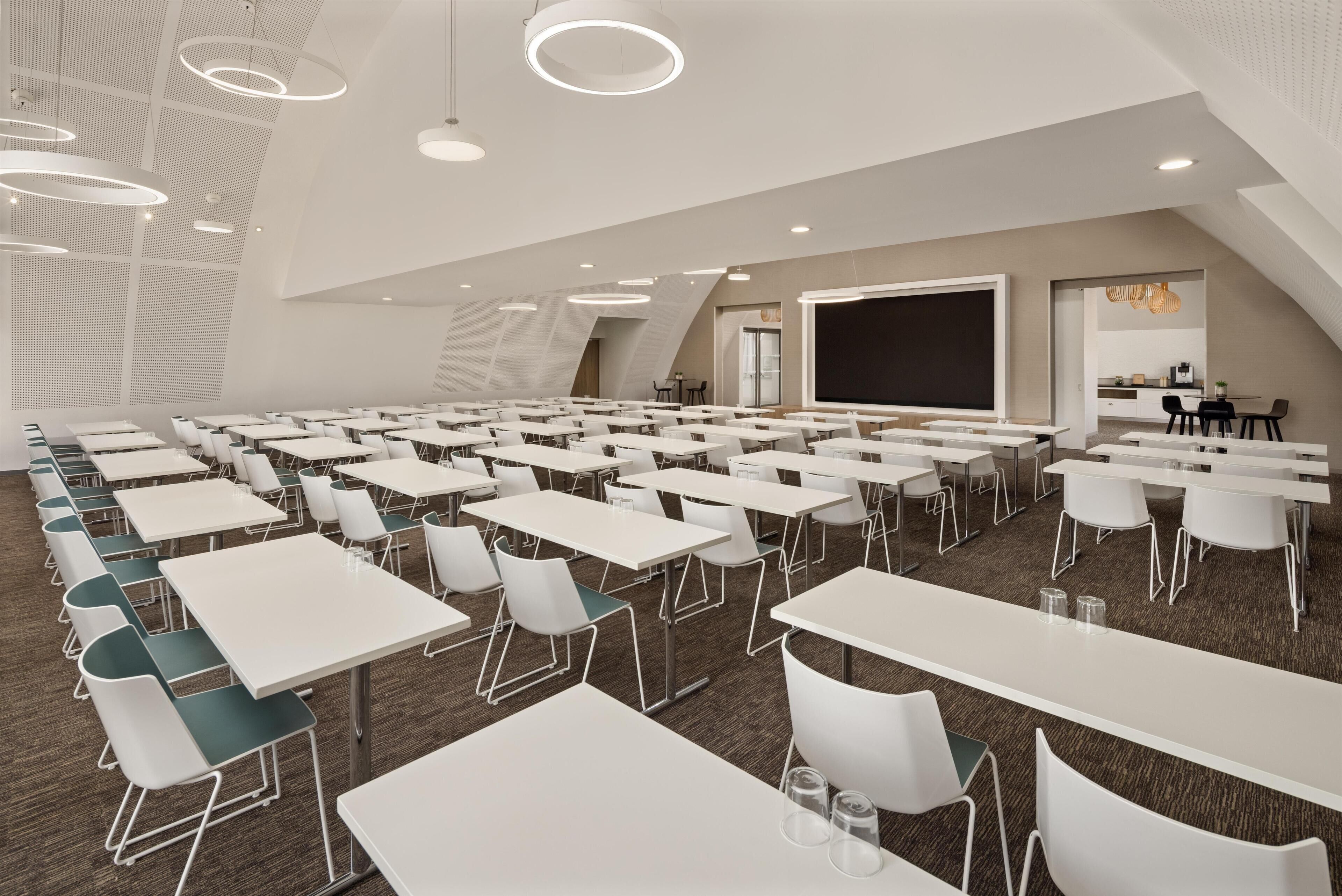 A conference room with white tables and chairs, a large screen, and a kitchen area in the background.