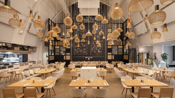 Un restaurant modern amb diverses taules i caires, llums penjants i una paret de quadre negre amb el menú.