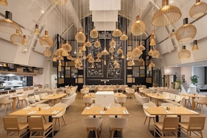 A modern restaurant with multiple dining tables and chairs, hanging pendant lights, and a chalkboard menu wall.