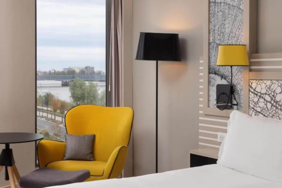 A hotel room with a bed, a yellow armchair, a small table, and a view of a river and bridge.