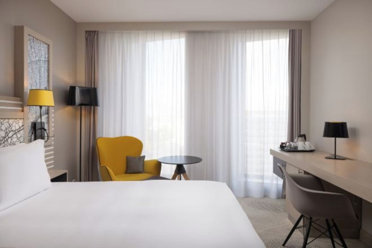 Hotelkamer met een bed, een gele fauteuil, een klein rond tafeltje, een bureau met stoel en een groot raam met dunne gordijnen.