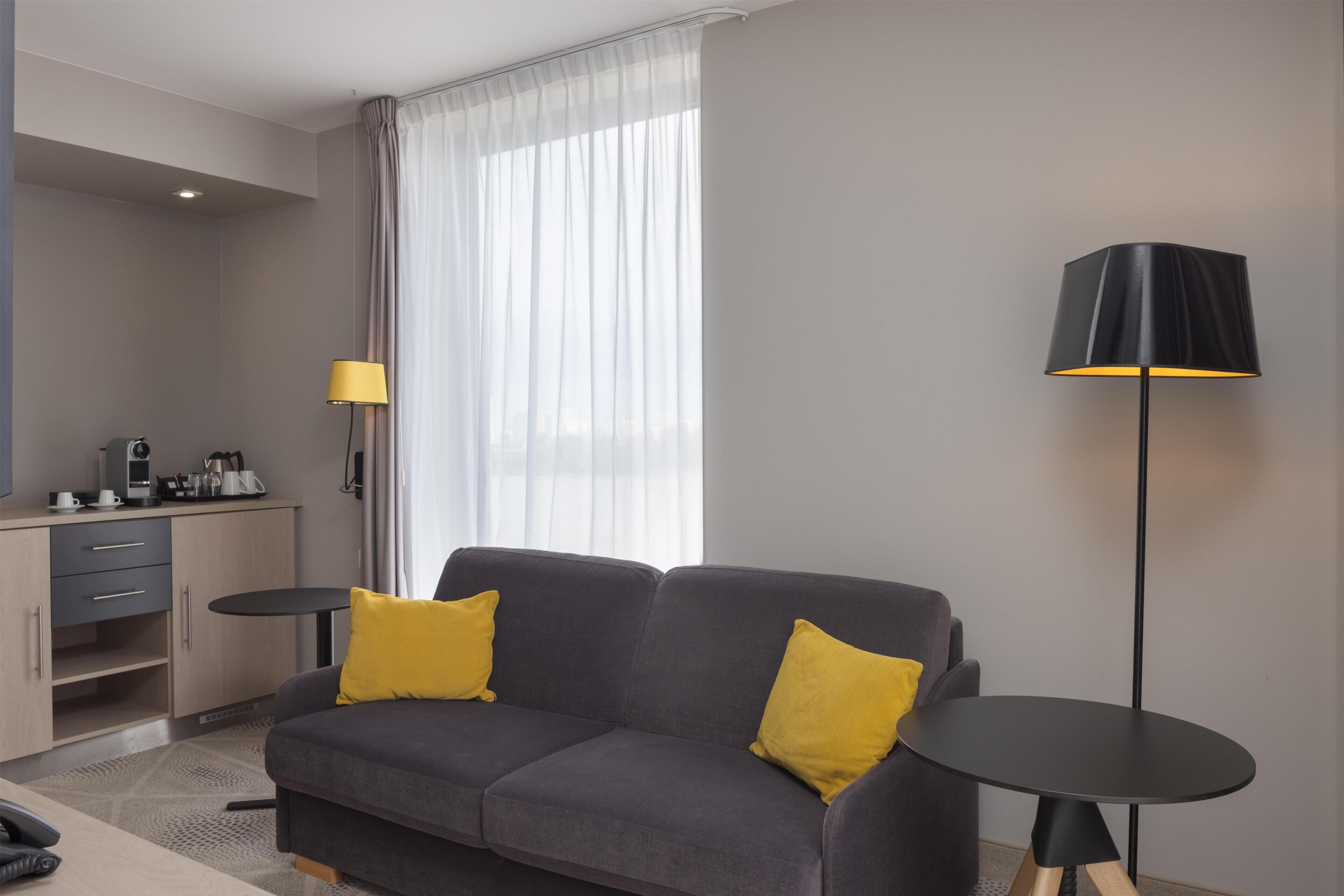 A modern hotel room with a grey sofa, two yellow cushions, a small round table, a black floor lamp, a desk with a coffee machine, and a large window with sheer curtains.