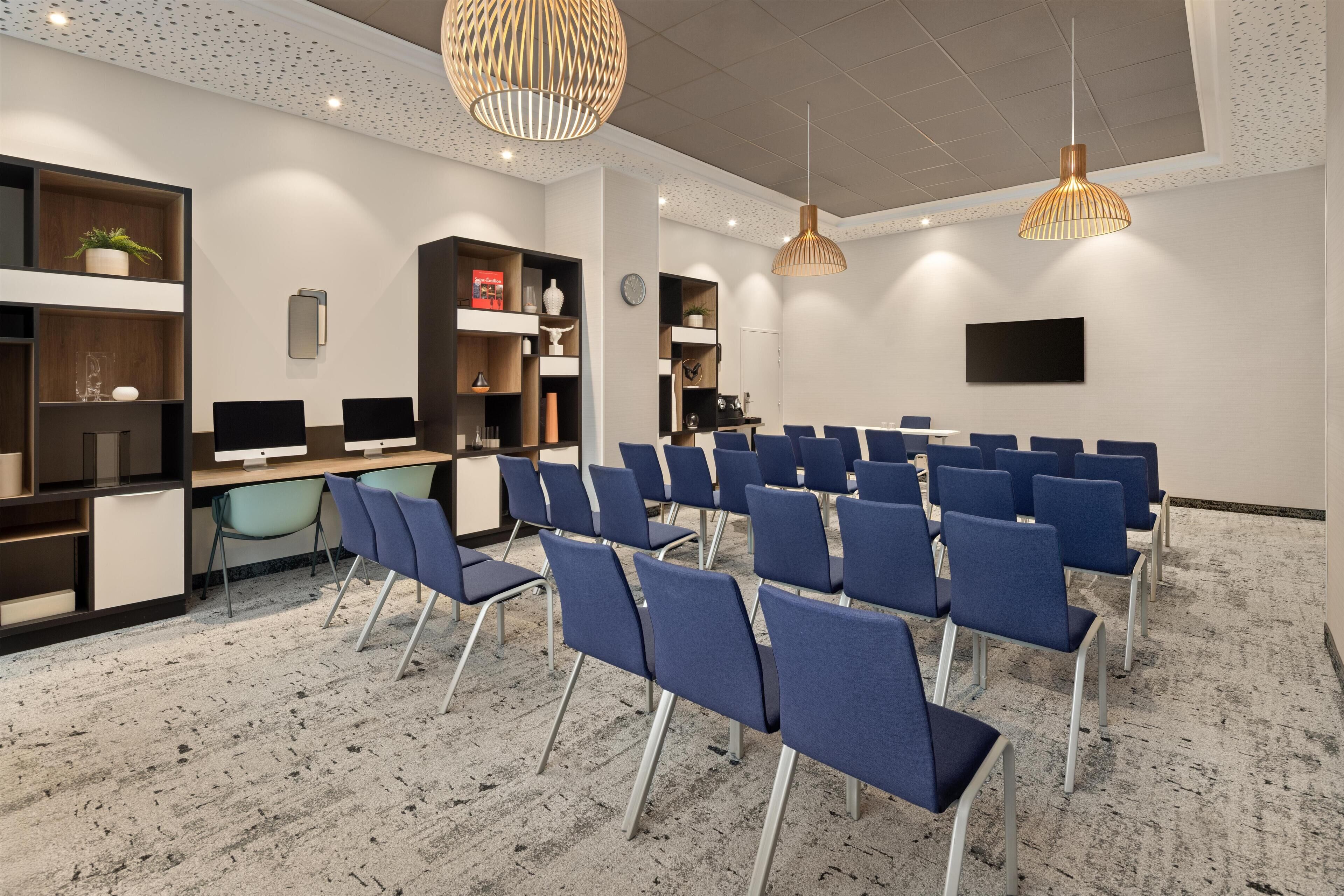 A conference room with rows of blue chairs, a wooden desk, shelves with various items, and pendant lighting.