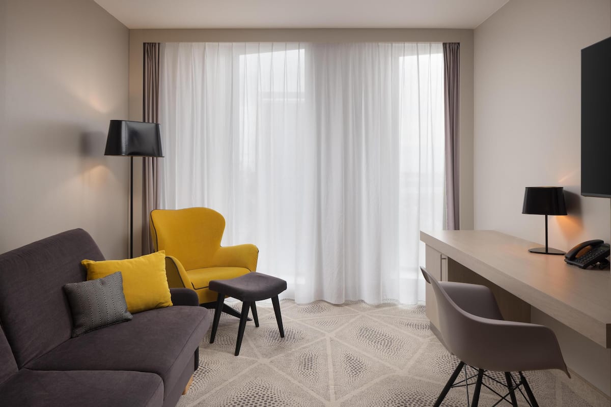 A hotel room with a grey sofa, a yellow armchair, a small table, and a TV on a stand.