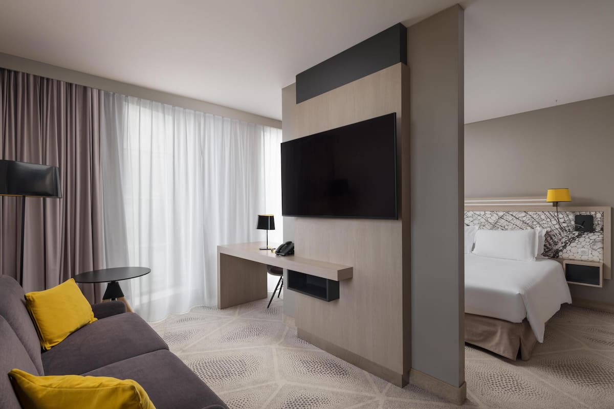 A modern hotel room with a sofa, a desk with a flat-screen TV, a bed, and a nightstand with a lamp.