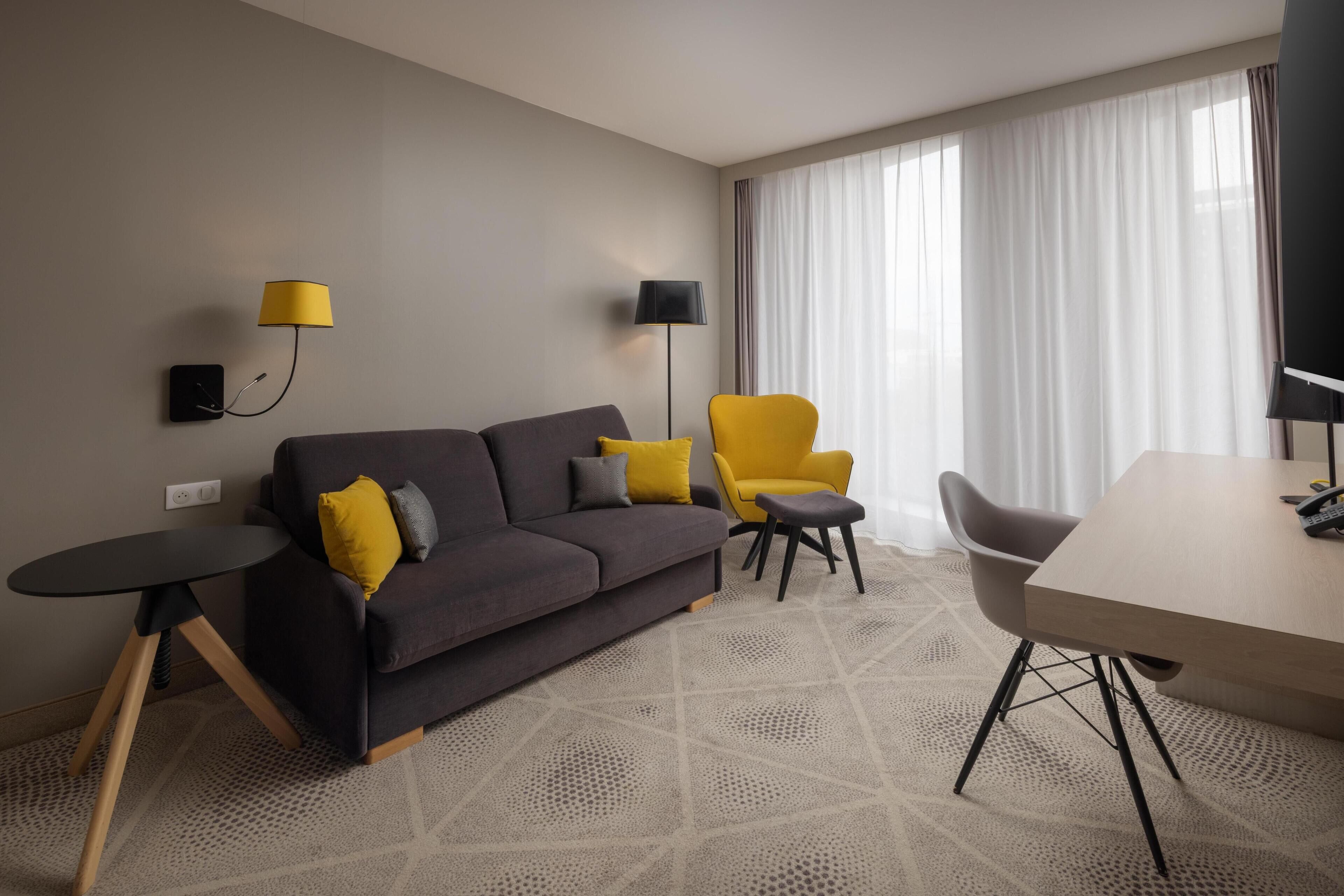 A modern hotel room with a dark grey sofa, a yellow armchair, a small round table, and a large window with sheer curtains.