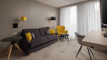 Une chambre d’hôtel moderne avec un canapé gris foncé, un fauteuil jaune, une petite table ronde et une grande fenêtre ornée de rideaux légers.