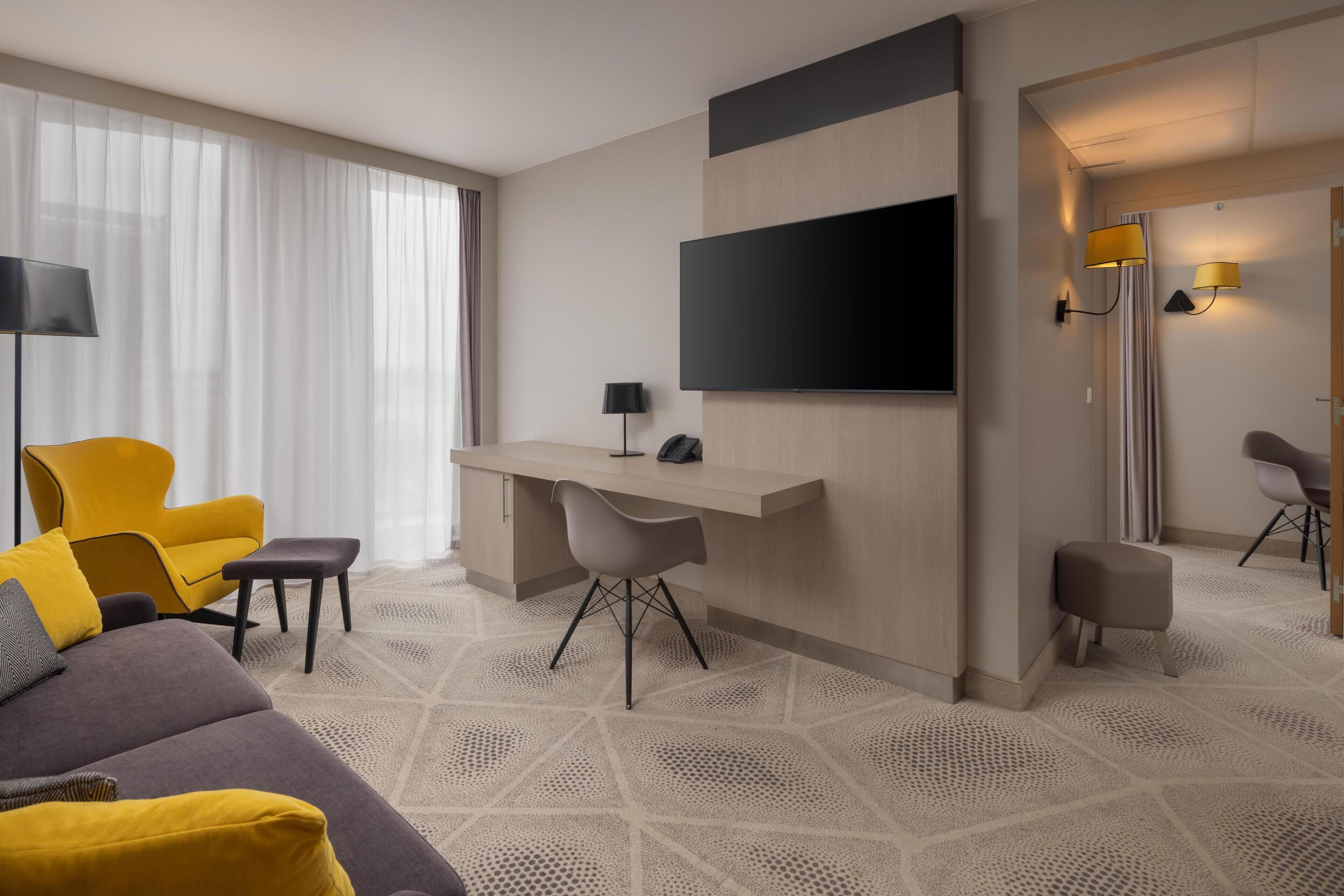 A modern hotel room with a sofa, a desk, a flat-screen TV, and a large window with sheer curtains.