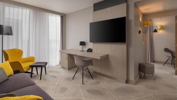 A modern hotel room with a sofa, a desk, a flat-screen TV, and a large window with sheer curtains.