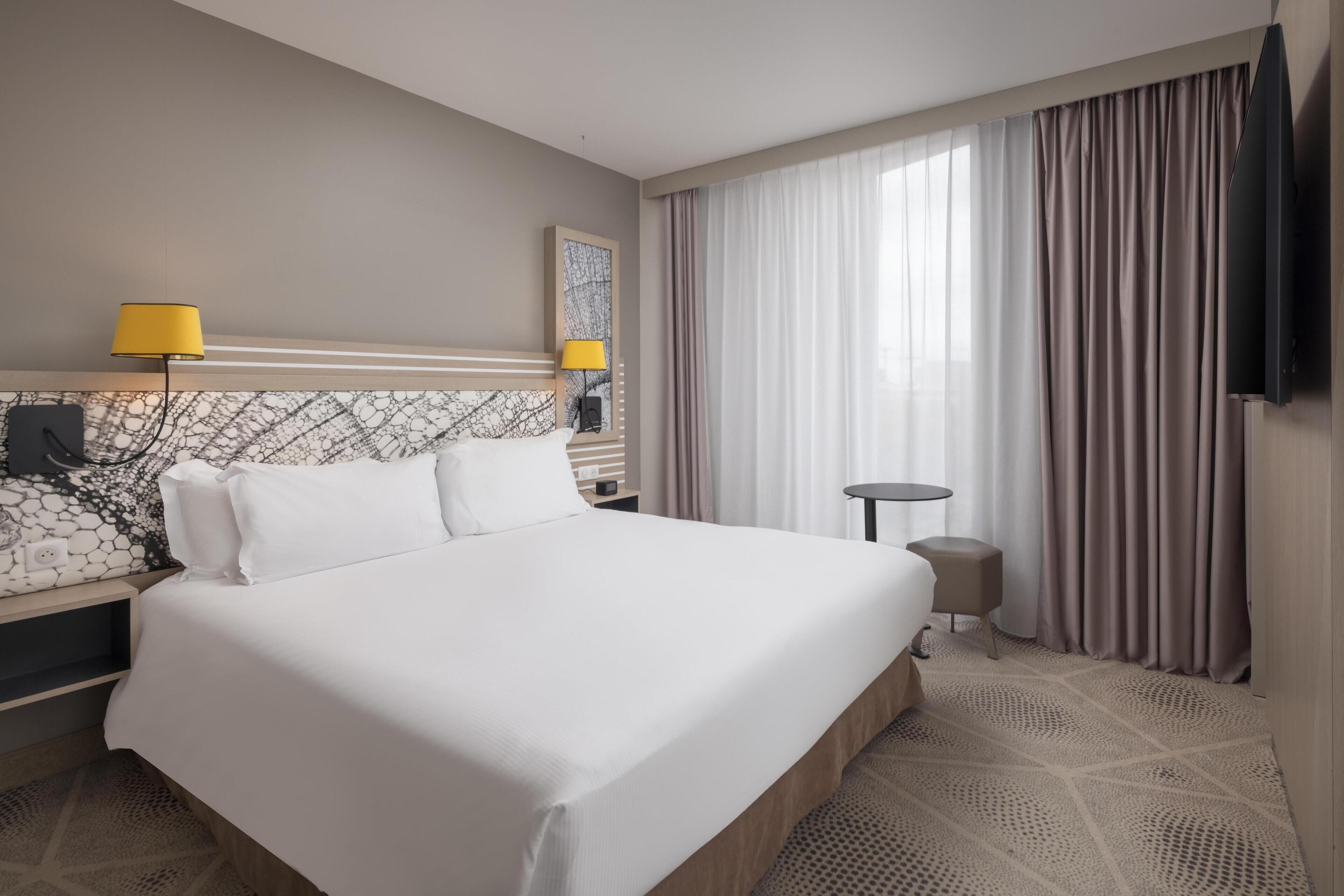 Habitación de hotel con una cama grande, una mesita de noche con lámpara, una pequeña mesa redonda y un televisor montado en la pared.