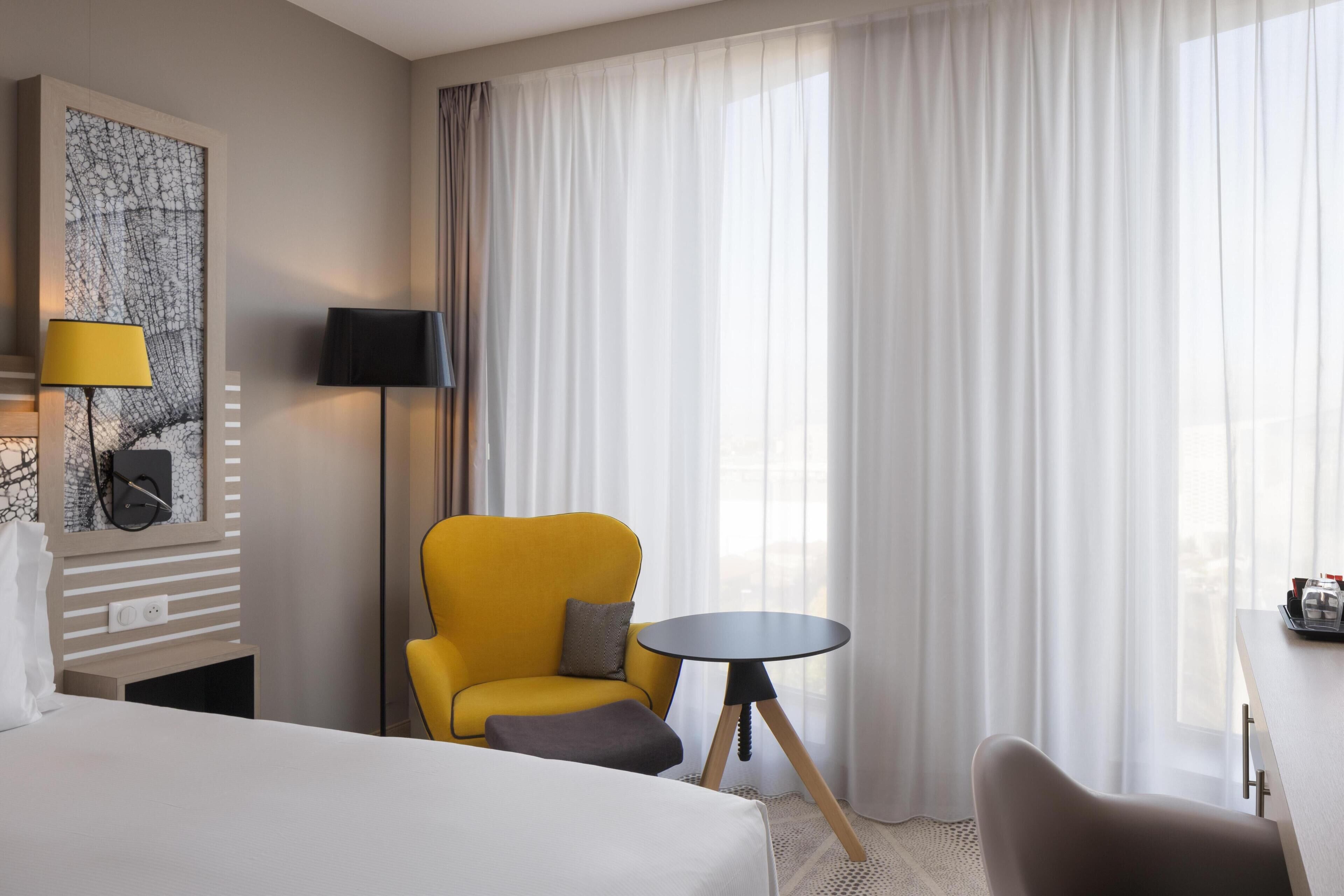 Une chambre d’hôtel avec un lit, un fauteuil jaune, une petite table ronde et une grande fenêtre avec des rideaux légers.