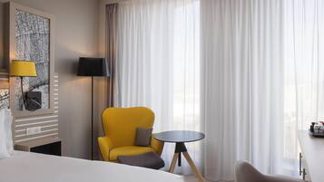 A hotel room with a bed, a yellow armchair, a small round table, and a large window with sheer curtains.
