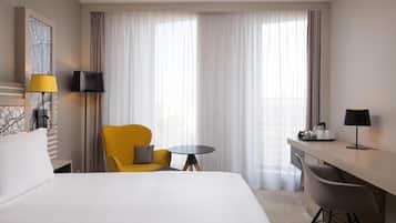 A hotel room with a large bed, a yellow armchair, a small round table, a desk with a chair, and a large window with sheer curtains.