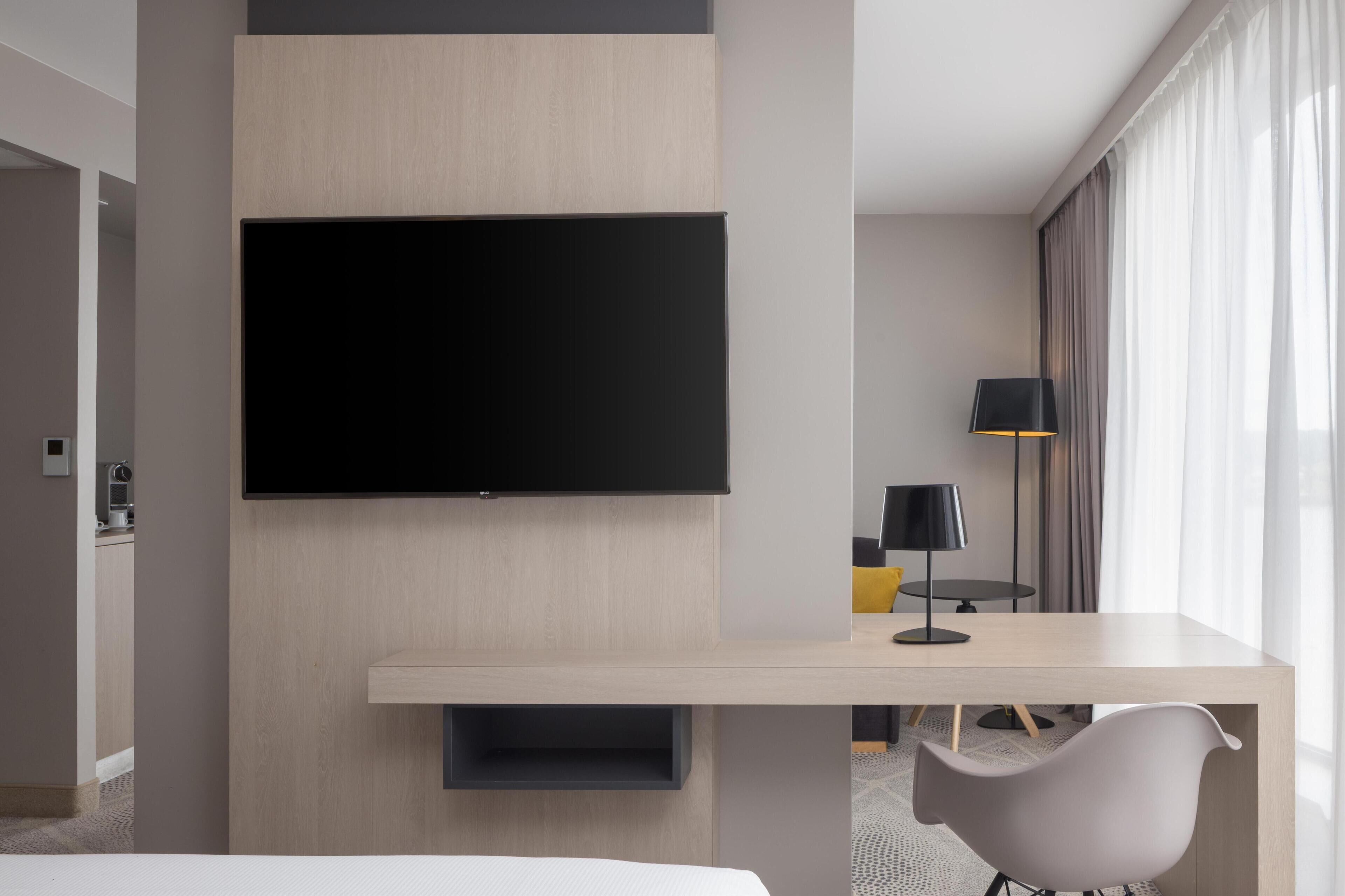 Ein modernes Hotelzimmer mit einem wandmontierten Flachbildfernseher, einem Holztisch, einem modernen Sessel und einer Lampe.