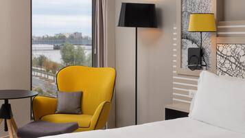 A hotel room with a large window, a yellow armchair, a black floor lamp, a white bed, and a small table.