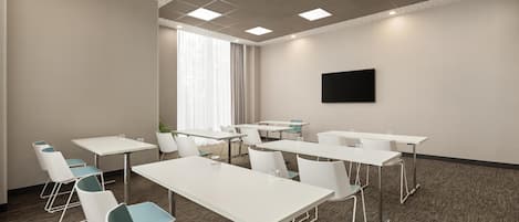 Un salón de conferencias con mesas y sillas blancas, un ventanal con cortinas y un televisor de pantalla plana en la pared.