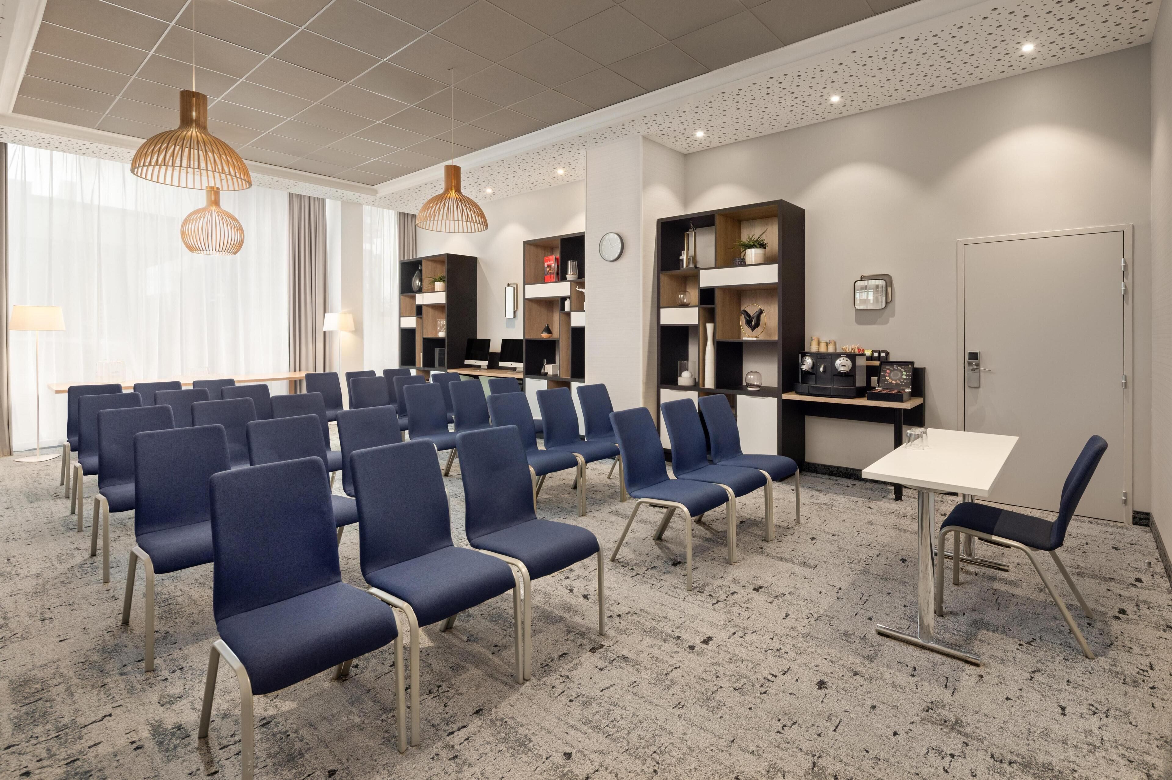 Una sala de conferències amb files de cadires blaves, una taula blanca i prestatges amb diversos objectes.