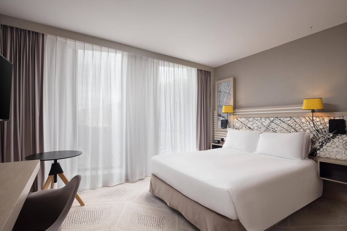 A hotel room with a bed, a desk, and a chair. There is a window with curtains, a lamp, and a wall-mounted TV.