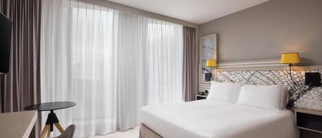 Een hotelkamer met een bed, een bureau en een stoel. Er is een raam met gordijnen, een lamp en een aan de muur bevestigde televisie.