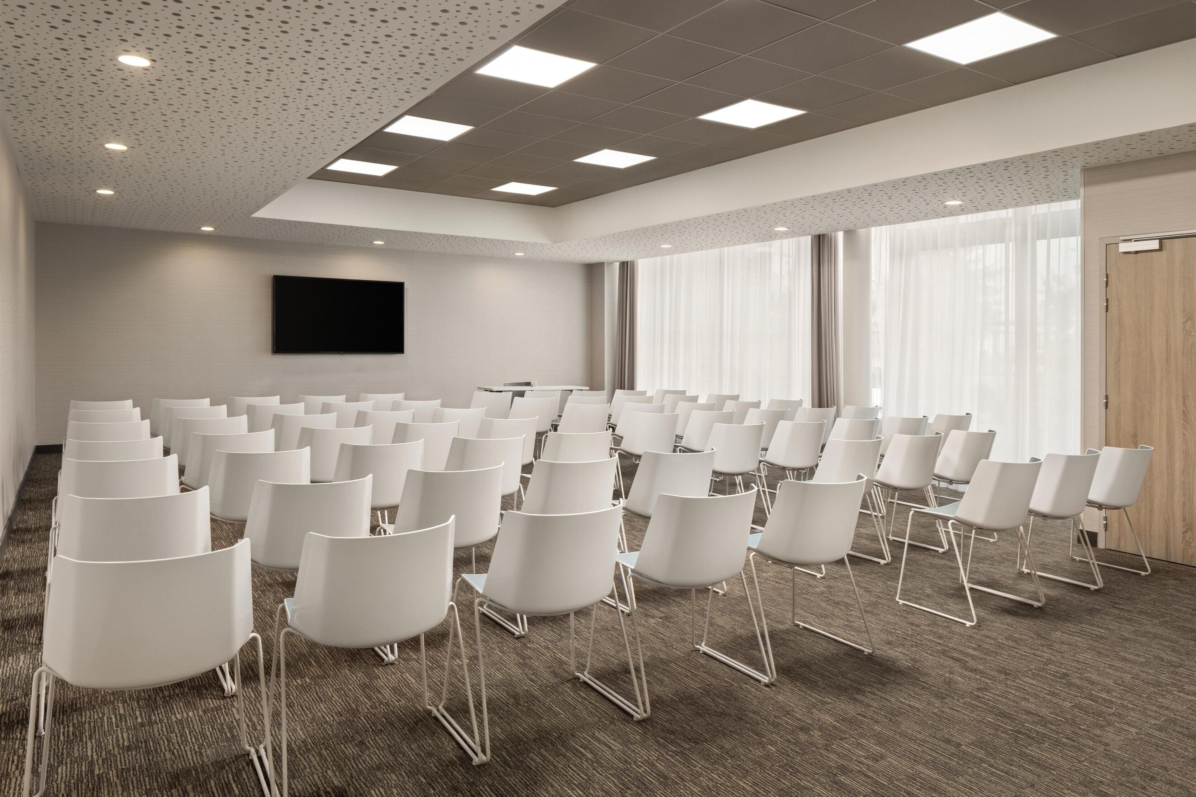 A conference room with rows of white chairs, a large flat-screen TV on the wall, and large windows with sheer curtains.