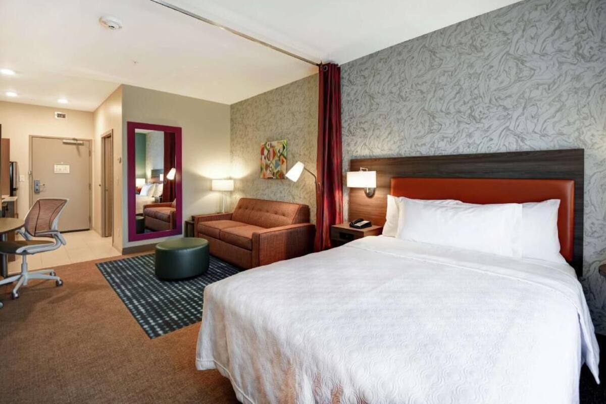 A hotel room with a large bed, a desk, a chair, a small ottoman, and a mirror.