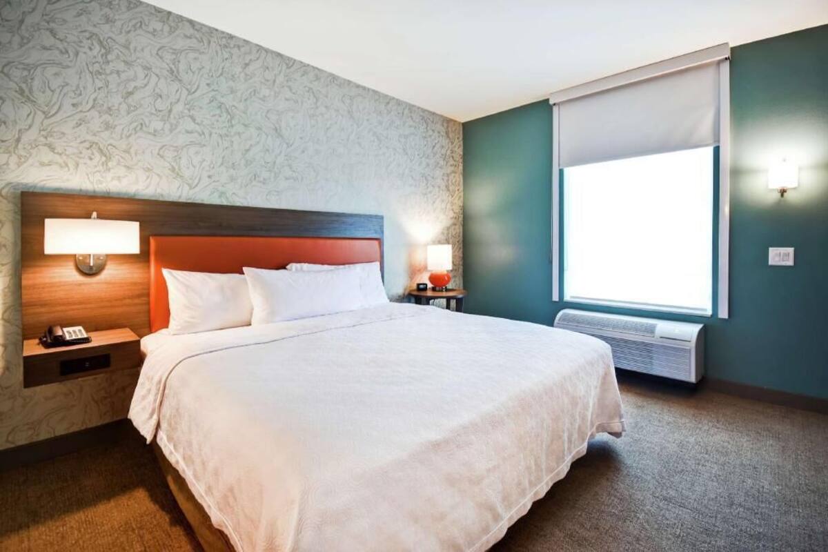 A hotel room with a bed, nightstands, lamps, a window, and an air conditioning unit.