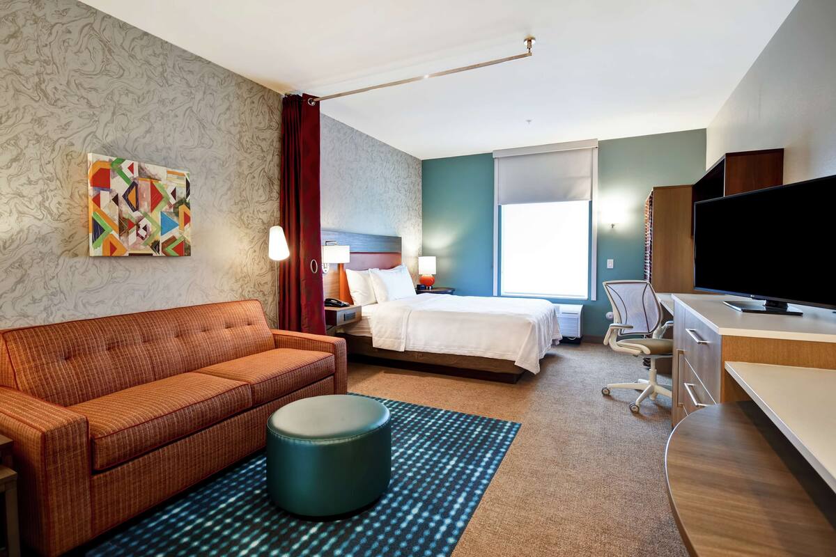 A hotel room with a bed, a sofa, a desk, a TV, and a painting on the wall.